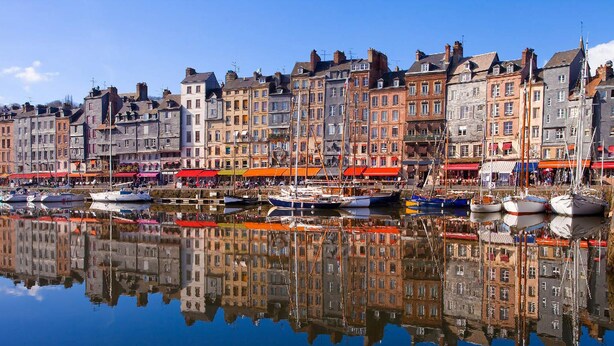 Un muelle con edificios históricos, un puerto deportivo con barcos y un reflejo claro en el agua.