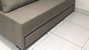 En soffa med en förvaringslåda, beige gardiner och en tavla med ett solnedgångsmotiv på väggen.