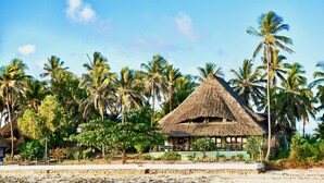 A thatched-roof hut with a veranda, surrounded by palm trees and a sandy beach. - THE PALMS VILLA - Six Bedroom Villa, Sleeps 12 (South East Zanzibar)