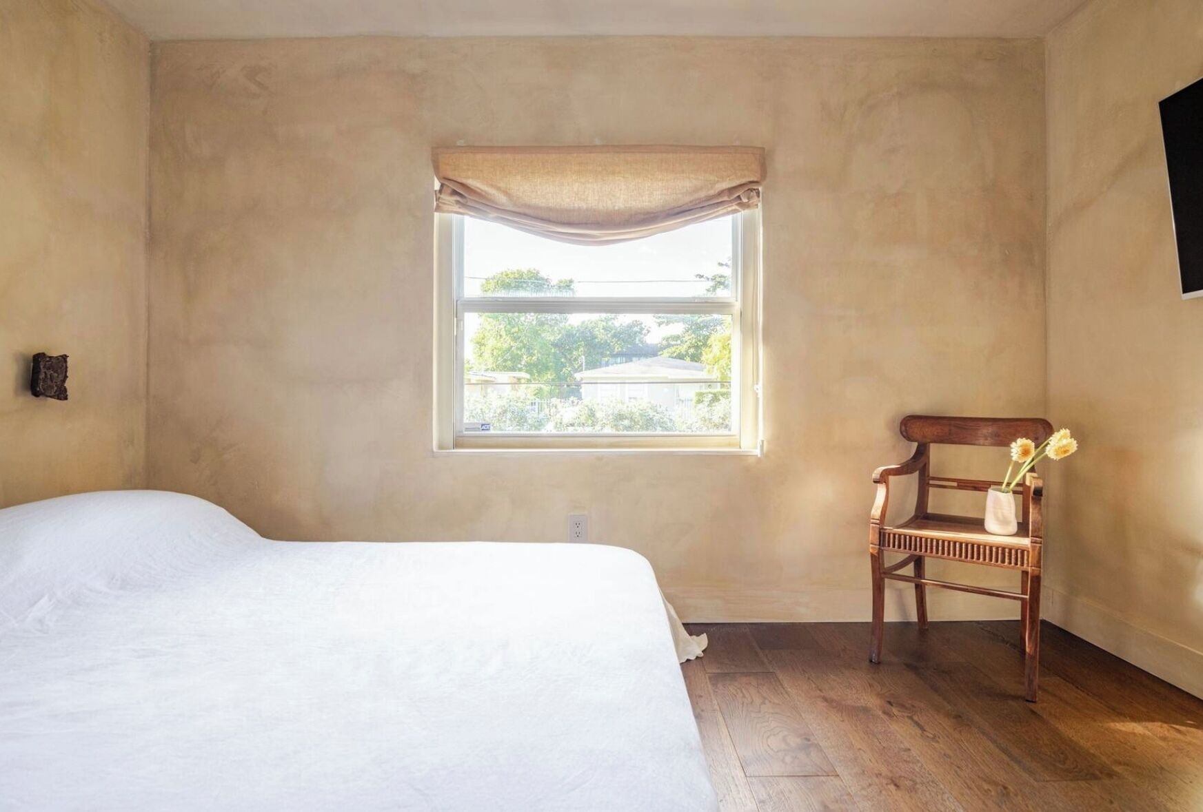 A bedroom with a single bed, a window with a curtain, a wooden chair with a vase of flowers, and a wall-mounted light switch.