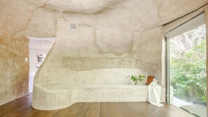 A room with a large, curved white bathtub, wooden flooring, and a view of greenery through a glass door.