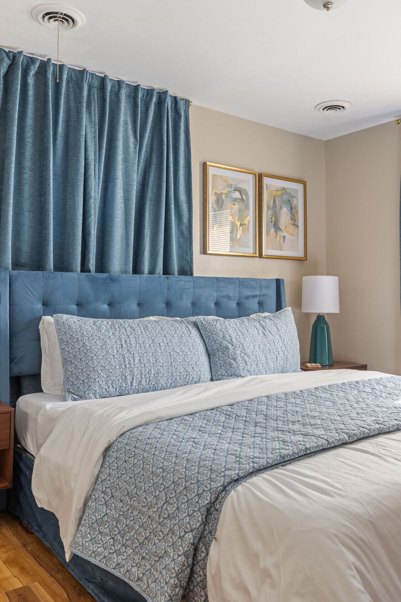 Une chambre à coucher avec une tête de lit capitonnée, un lit recouvert d’une literie à motifs, une table de chevet avec une lampe et deux œuvres d’art encadrées accrochées au mur.