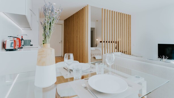 A modern dining area with a glass table set for four, white plates, and clear glasses. A wooden partition separates the dining area from a living space. - VIVA Formosa Lofts 1 (Porto)