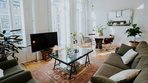 Ein geräumiges Wohnzimmer mit einem großen Flachbildfernseher, einem Glastisch und einer gemütlichen Couch. Mehrere Zimmerpflanzen und eine Essecke mit Tisch und Stühlen sind ebenfalls vorhanden.