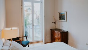 Ein Schlafzimmer mit Bett, Stuhl, Schminktisch und einem Fenster mit Blick auf Gebäude.