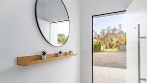 Un interior moderno con un estante de madera con plantas pequeñas, un espejo grande y una puerta que da a un espacio exterior con un camino de grava y árboles.