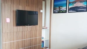 Ett rum med en tv som är monterad på en väggpanel av trä, en bokhylla och en tavla som föreställer en strandscen.