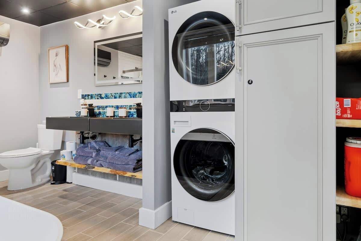 A modern laundry room with a front-loading washer and dryer, a toilet, and a shelf with cleaning supplies.