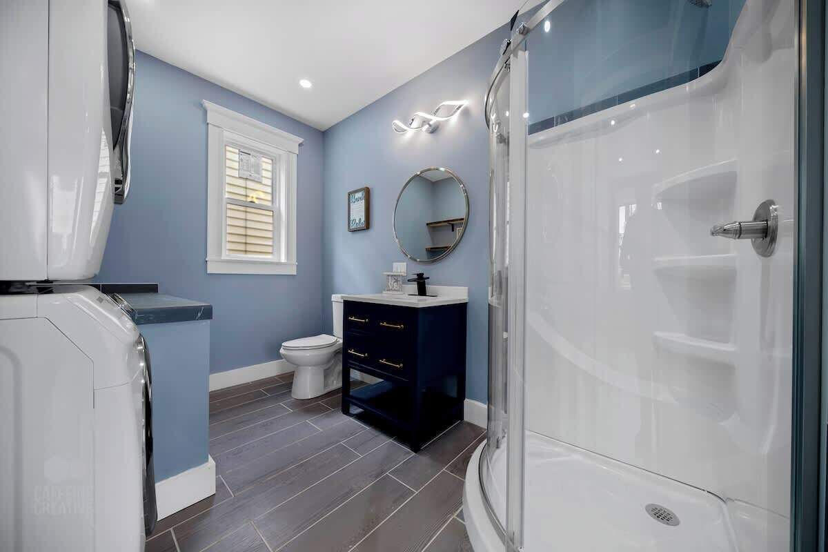 A bathroom with a washing machine, a toilet, a dark blue vanity, a round mirror, and a glass shower enclosure.