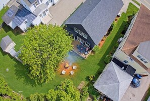 An aerial view of a residential area with a large tree, a house with a dark roof, and a visible fire pit.