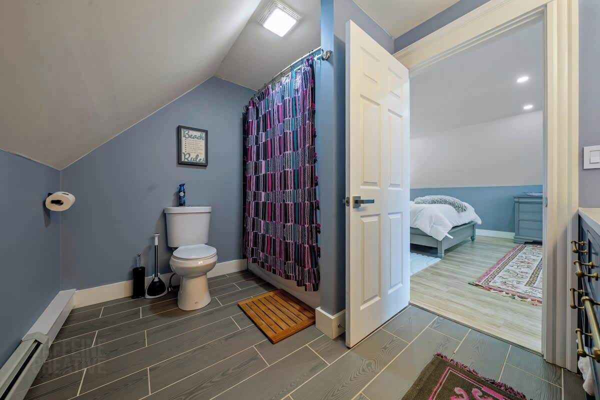 A bathroom with a toilet, shower curtain, and a door leading to a bedroom with a bed and a nightstand.