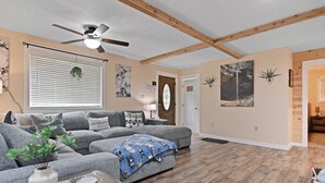 Un salon avec un canapé gris, un ventilateur de plafond, des poutres en bois, un plancher en bois et une porte ornée d’un panneau d’avertissement.