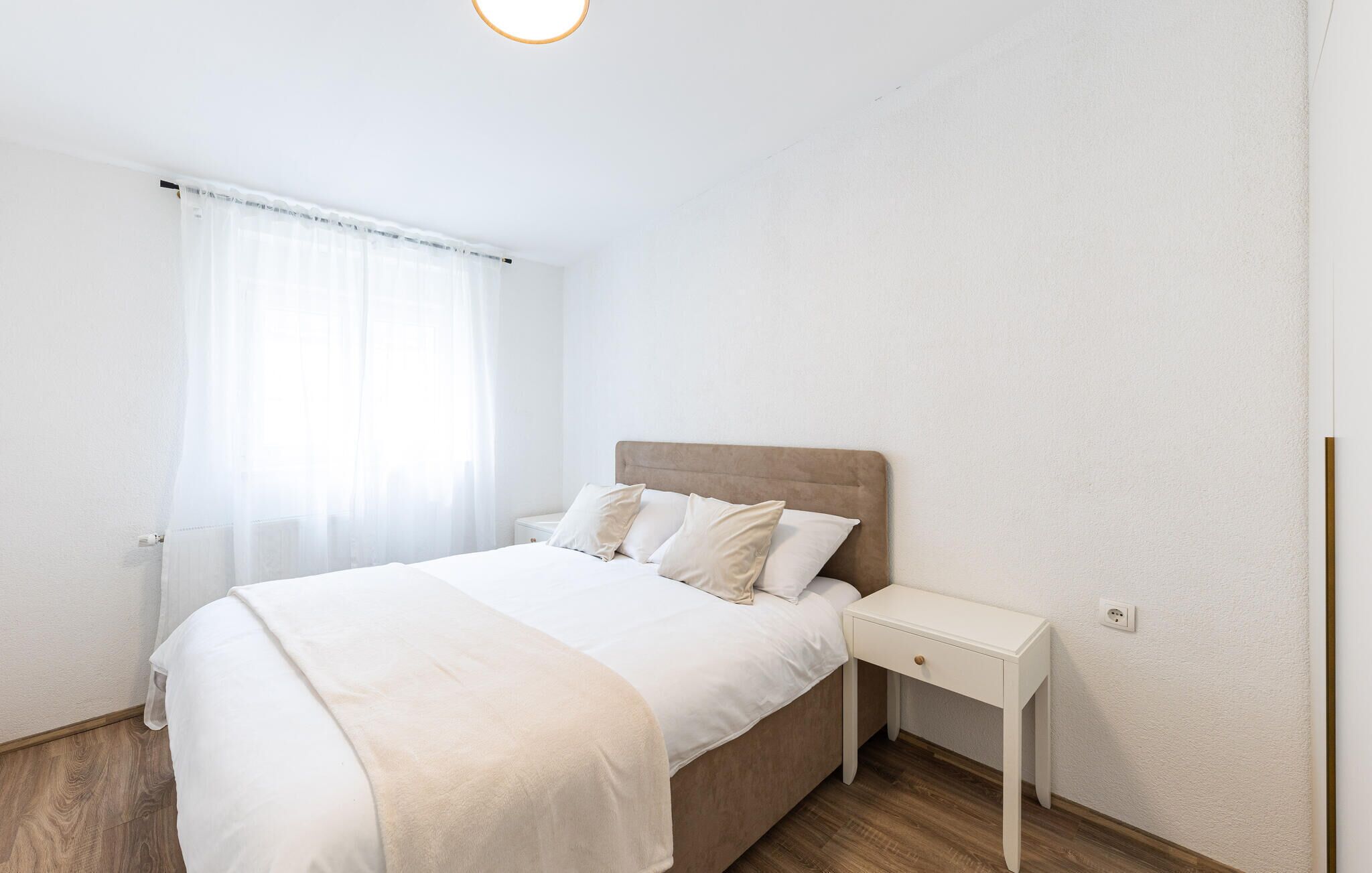 A minimalist bedroom with a large bed, a bedside table, and a window with sheer curtains.