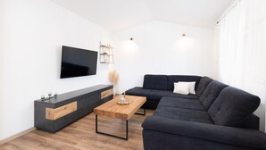 A modern living room with a dark grey sofa, a wooden coffee table, a TV mounted on the wall, and a minimalist shelving unit. - Stunning home in Sinj (Sinj)