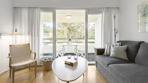 En moderne stue med en grå sofa, et hvidt sofabord, en fletstol og en balkon med udsigt over træer og bygninger.