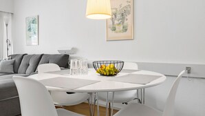 Et moderne spiseområde med et rundt hvidt bord, hvide stole, en sort frugtskål i metal og en hængende lampe.