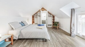 Un dormitorio con techo inclinado, una cama con colchón blanco y almohadas grises, una silla de madera, una mesita pequeña y un armario con las puertas abiertas.
