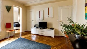 A living room with a wooden floor, a white entertainment unit with a TV, a red chair, and a large potted plant. - Manu apartment   Paris (Paris)