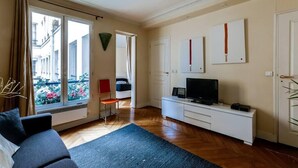 A modern living room with a sofa, a TV on a white cabinet, and a view of a balcony with flowers. - Manu apartment   Paris (Paris)