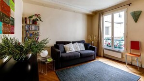 A living room with a dark sofa, a bookshelf, a potted plant, and a window with a view of a balcony. - Manu apartment   Paris (Paris)