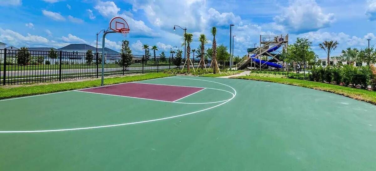 Un terrain de basket en plein air, avec une surface de jeu rouge et verte, entouré d’une clôture et de palmiers.