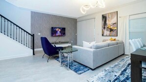 A modern living room with a grey sofa, a blue chair, a glass coffee table, and a TV mounted on the wall. - Go!#05 - CHAMPIONS GATE (Kissimmee)