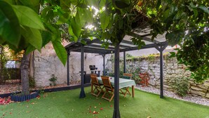 Un patio techado al aire libre con una mesa y sillas, rodeado por un muro de piedra y vegetación.