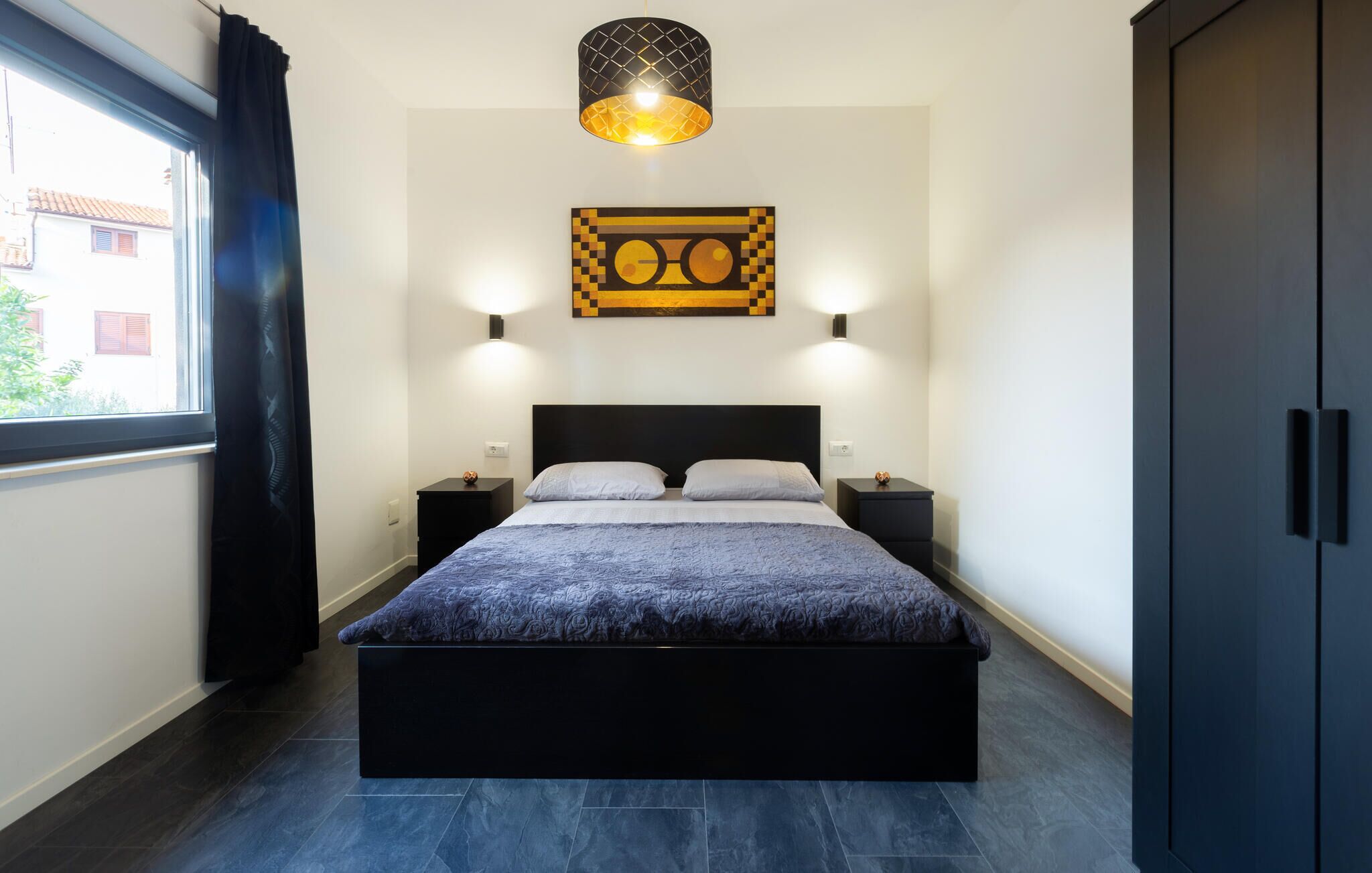 Une chambre à coucher comprenant un lit, deux tables de chevet, une œuvre d’art accrochée au mur et une fenêtre avec des rideaux.