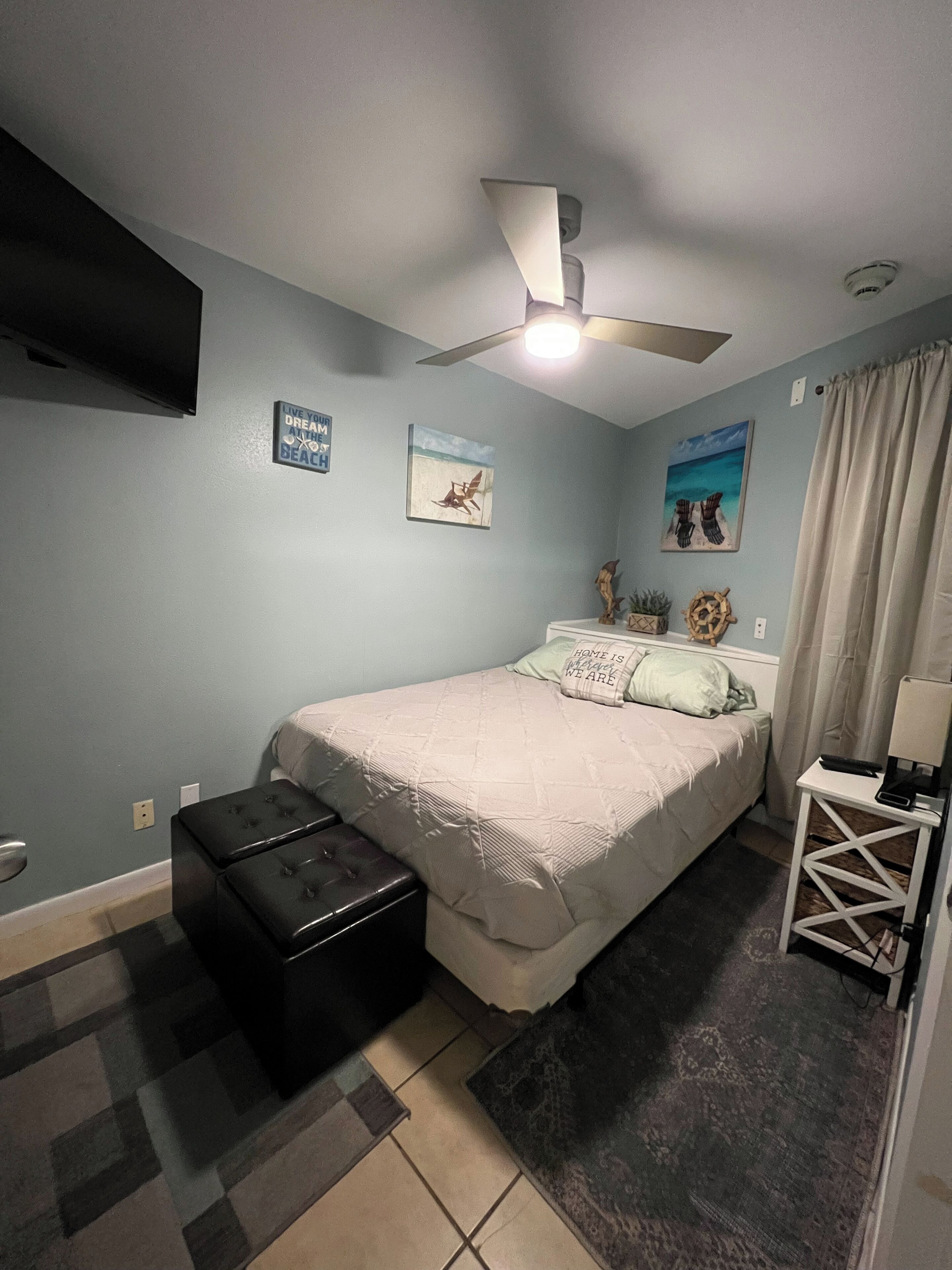 Ein Schlafzimmer mit einem Bett, einem Deckenventilator, einem an der Wand befestigten Fernseher und einem kleinen Tisch mit einer Lampe.