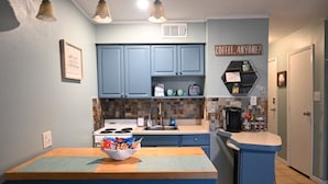 A kitchen with blue cabinets, a central island with a bowl of snacks, a coffee machine, and a sign that says "Coffee, anyone?".