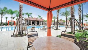 An outdoor pool area with lounge chairs and a table under a red canopy, surrounded by palm trees and a building in the background.