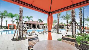 An outdoor pool area with lounge chairs and a table under a red canopy, surrounded by palm trees and a building in the background.