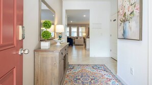 Een gang met een houten dressoir, een spiegel, een lamp, een vaas met bloemen en een kamerkleed met een patroon.
