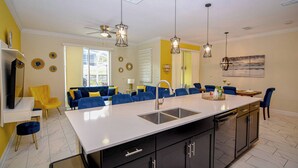 Cozinha moderna com ilha central, armários pretos e bancada branca. Possui área de jantar com cadeiras azuis e sofá amarelo.