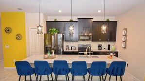 Uma cozinha moderna com armários escuros, uma ilha central com banquetas azuis, eletrodomésticos de aço inoxidável e iluminação pendente.