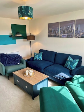 A modern living room with a blue sofa, a matching armchair, a coffee table, a floor lamp, and a cityscape painting.