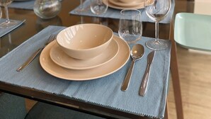 Una mesa de comedor preparada con platos, tazones, cubiertos y copas de vino, con un reloj en la pared al fondo.
