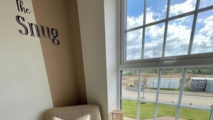Una habitación de esquina con un sillón estampado, una ventana con vista a un estacionamiento y una pared con el texto 'The Snug'.