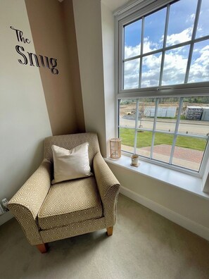 A corner room with a patterned armchair, a window with a view of a parking lot, and a wall with 'The Snug' text.