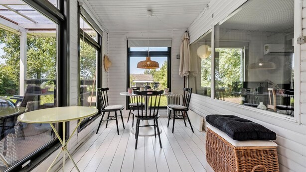 Een zonnig terras met een ronde tafel en stoelen, een rieten bank en grote ramen met uitzicht op de omgeving.