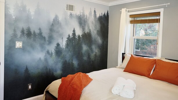 A bedroom with a forest mural, a ceiling fan, a window with curtains, and a neatly made bed with orange and white pillows. - Luxurious 3-Level Home Minutes from Washington D.C. (Arlington)