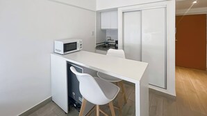 Une petite cuisine équipée d’un four à micro-ondes, d’un évier et d’un petit plan de travail. On y trouve une chaise blanche et un meuble avec une porte coulissante.