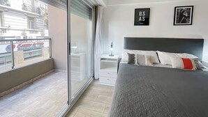 Une chambre moderne avec un grand lit, une table de chevet, une commode et une vue sur le paysage urbain, visible à travers une porte-fenêtre coulissante.