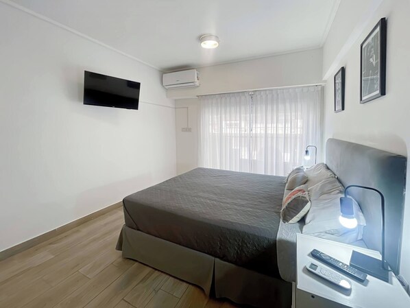 A bedroom with a bed, a bedside table with a lamp, a TV mounted on the wall, and an air conditioning unit. - Modern Studio With Amenities in the Heart of the City -u12 (Buenos Aires)