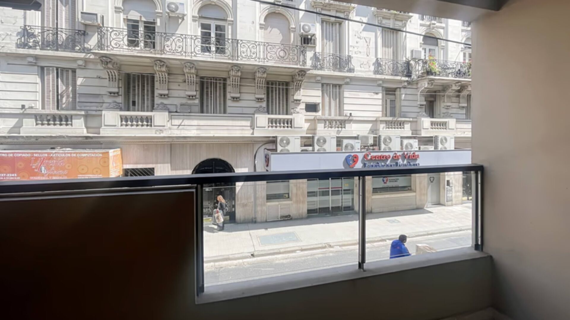 A view from a window overlooking a street with a building that has ornate balconies and air conditioning units.