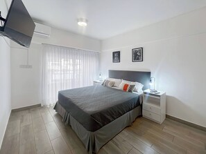 Ein modernes Schlafzimmer mit einem großen Bett, Nachttischen, einem Flachbildfernseher und einem Fenster mit leicht transparenten Vorhängen.