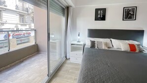 Quarto com cama, mesa de cabeceira, janela com vista para um edifício e duas fotos emolduradas na parede.