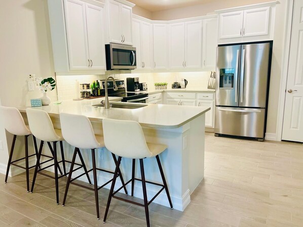 A modern kitchen with white cabinetry, a stainless steel refrigerator, a microwave, and a bar stool area.