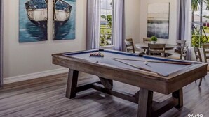 Une table de billard dans une pièce ornée de deux tableaux représentant des bateaux, avec un ventilateur de plafond et un coin repas comprenant des chaises et une table.