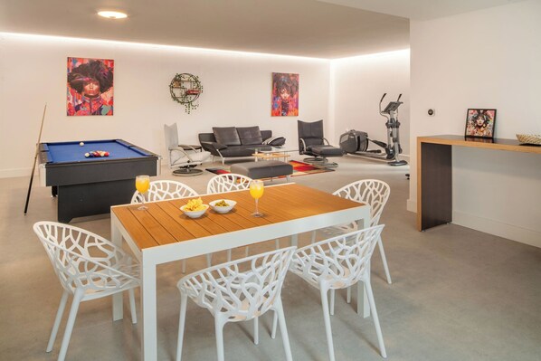 Una sala de estar moderna con mesa de billar, sillas blancas, mesa de comedor de madera y una cinta de correr.